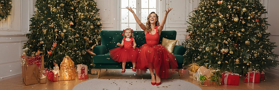 Mom and toddler wearing matching red bow-shoulder bodysuits on a green sofa between Christmas trees, surrounded by gold confetti. A joyful, elegant holiday moment from Motch’s mommy and me collection.