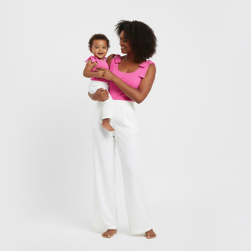 Mom holding her baby while wearing matching Hot Pink bow-shoulder bodysuits from Motch, paired with white pants, capturing the elegance and joy of coordinated mommy-and-me outfits.