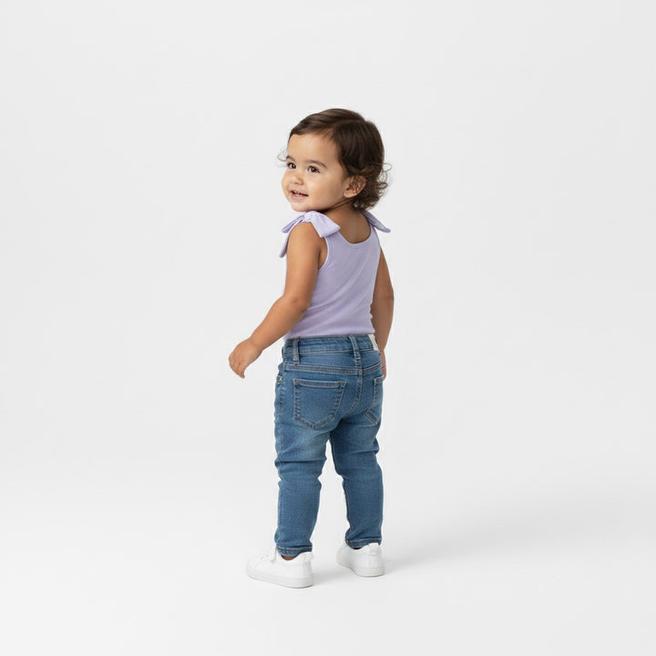 Toddler turning and smiling in a Motch Lavender bow-shoulder bodysuit and jeans — back view showing the smooth scoop neckline and playful bow detail from the mommy-and-me matching collection.
