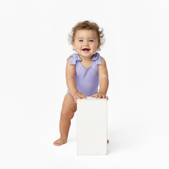 Baby standing and smiling while holding a white block in a Motch Lavender bow-shoulder bodysuit — soft, stretchy, and made for everyday comfort and sweet mommy-and-me matching moments.