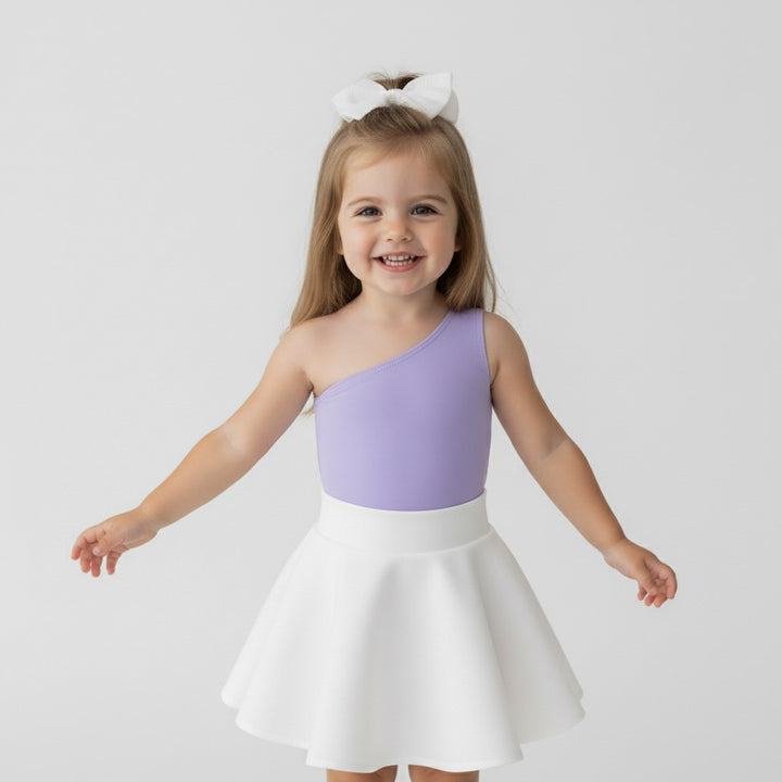 Toddler wearing a Motch Lavender one-shoulder bodysuit with a white skirt and bow, smiling playfully — part of the matching mommy-and-me collection designed for everyday comfort and photo-worthy moments.