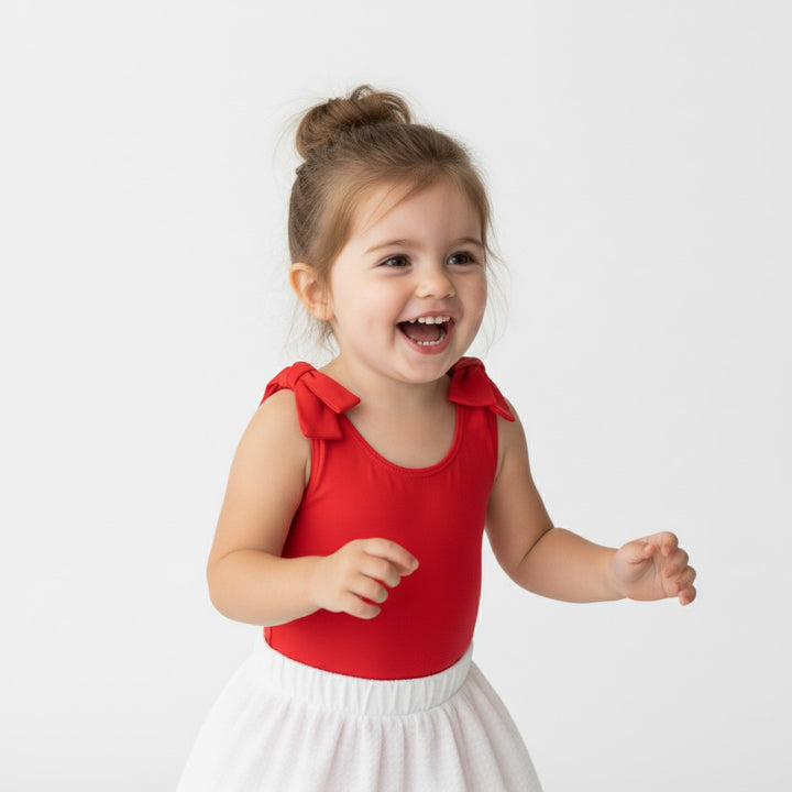 Smiling toddler girl in a red Motch bow-shoulder bodysuit and white skirt, captured mid-laughter — a cheerful mommy-and-me matching look for holidays or family photos.