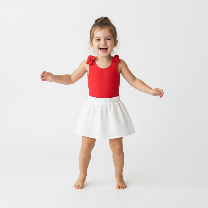 Toddler girl laughing and dancing in a red Motch bow-shoulder bodysuit with a white skirt — a bright, joyful look perfect for holidays, photos, or everyday play.