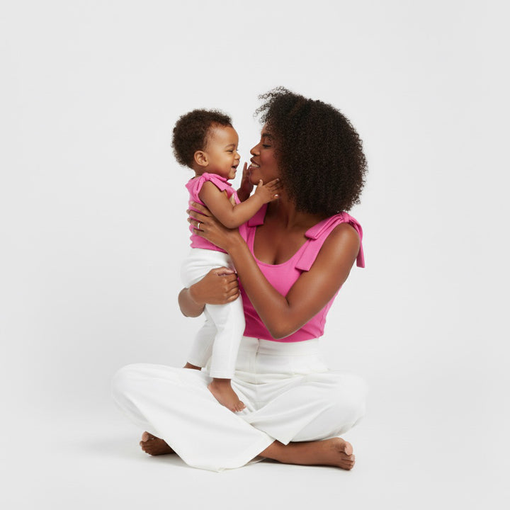 Mom and baby smiling and cuddling in matching hot pink Motch bow-shoulder bodysuits with white pants — a sweet mommy-and-me moment that captures connection, comfort, and color.