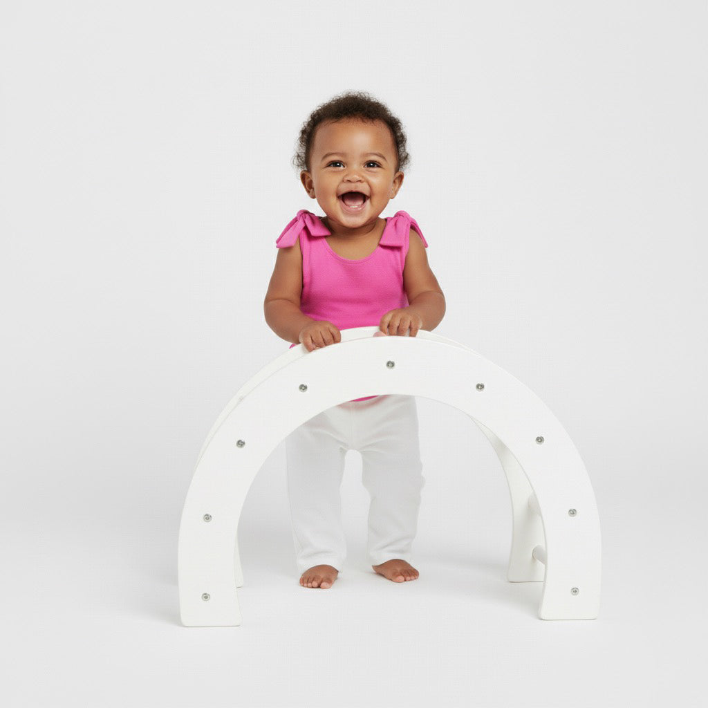Laughing baby standing and holding a white play arch in a bright pink Motch bow-shoulder bodysuit with white pants — soft, playful, and made for comfortable all-day movement.