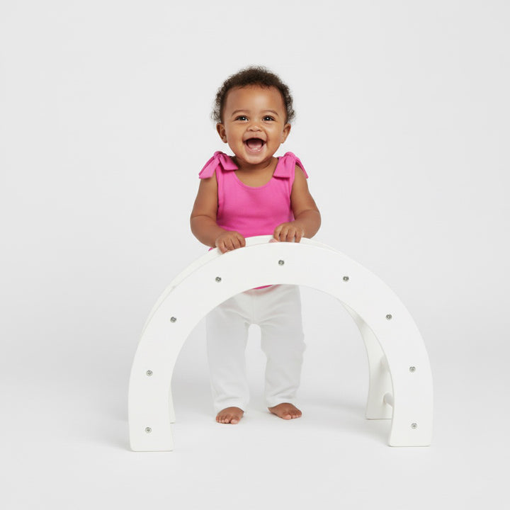Laughing baby standing and holding a white play arch in a bright pink Motch bow-shoulder bodysuit with white pants — soft, playful, and made for comfortable all-day movement.