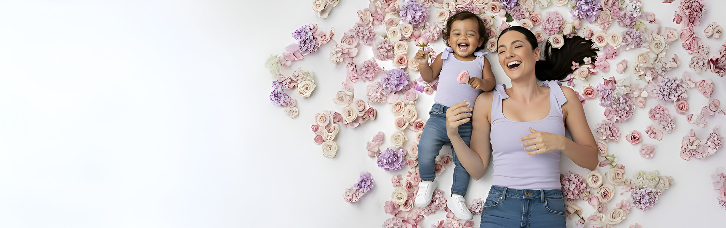 Mom and toddler lying on a white background surrounded by pastel flowers, laughing in matching lavender Motch bow-shoulder tops and jeans — joyful mommy-and-me moment celebrating connection and comfort.