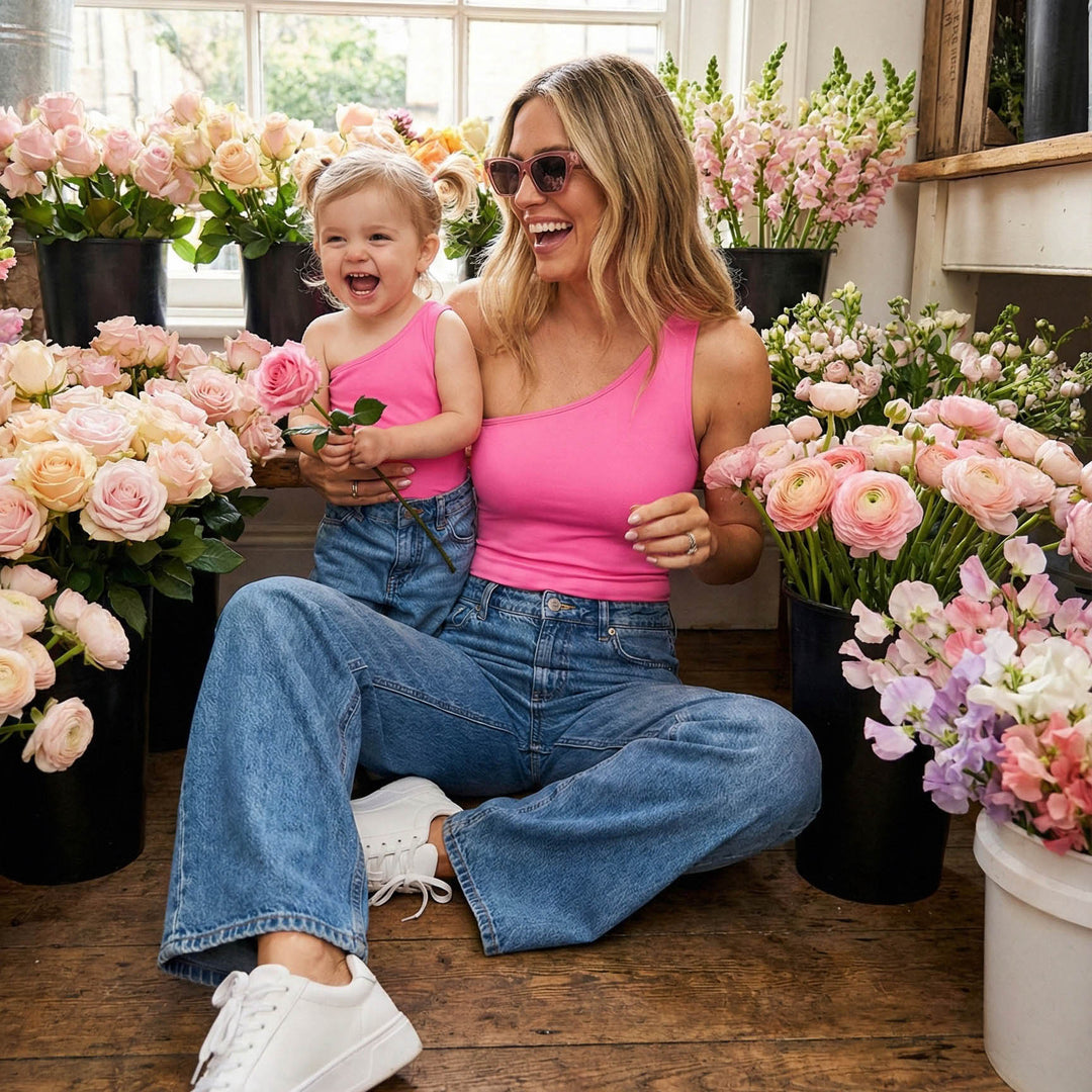 Mother and toddler wearing matching pink one-shoulder bodysuits and jeans, sitting together in a flower shop surrounded by blooms